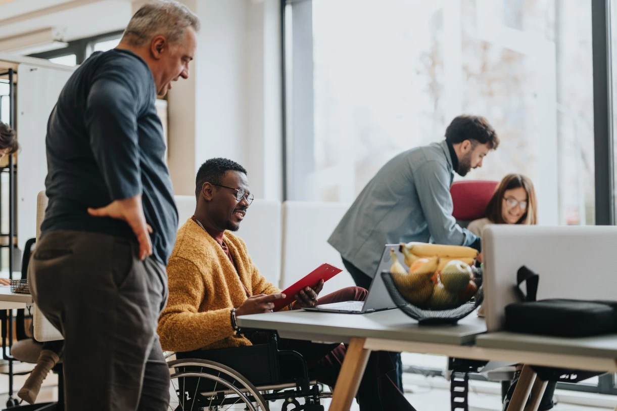 Smiling disabled man in wheelchair sitting at desk while looking at clipboard and talking to colleague