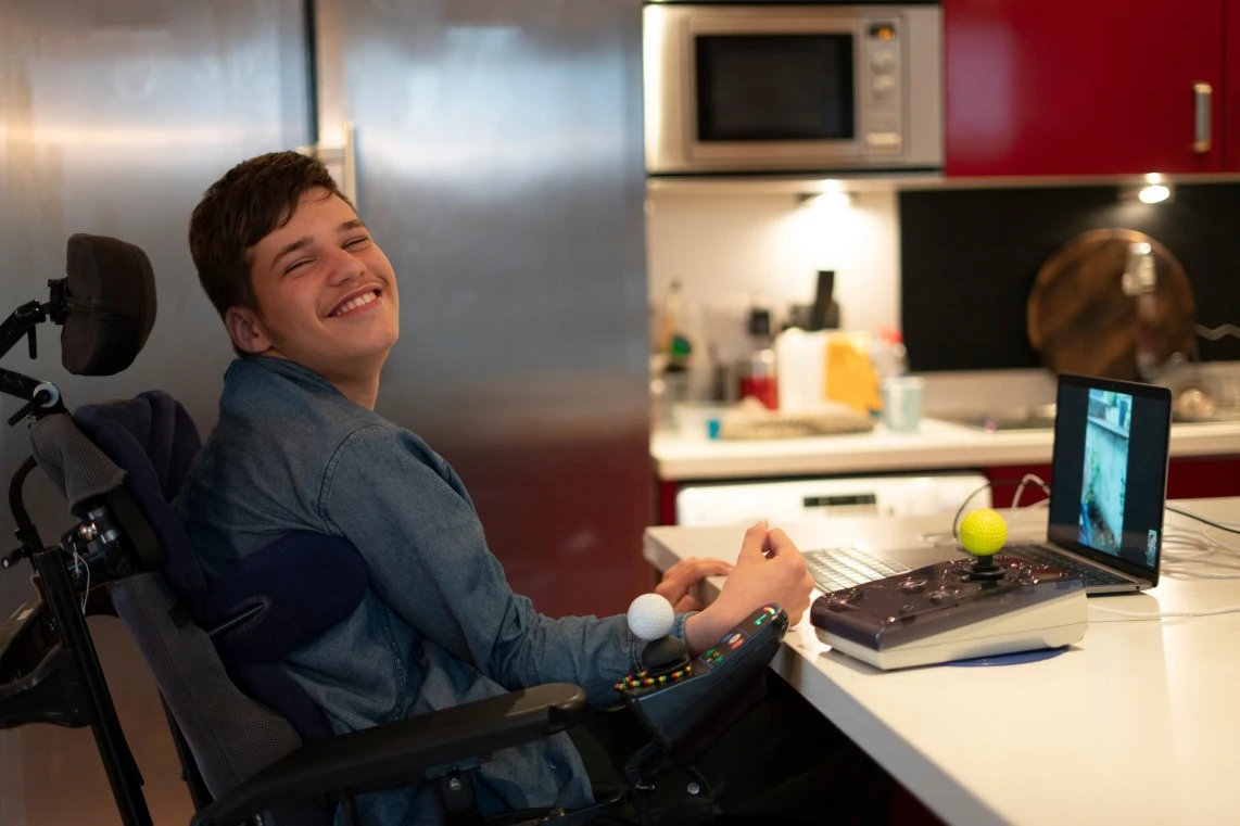 Disabled man in wheelchair smiling at camera while sitting at kitchen bench with his laptop